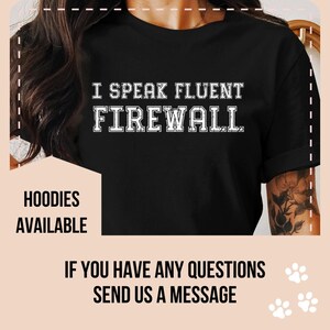May include: Black t-shirt with white text that reads "I speak fluent firewall". The text is in a vintage style font. The shirt is being modelled by a person with tattoos on their arm. The text "Hoodies available" is at the bottom left of the image. The text "If you have any questions send us a message" is at the bottom of the image.