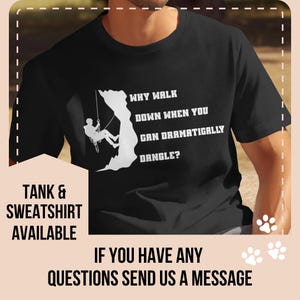 May include: Black t-shirt with a white graphic of a rock climber and the text "WHY WALK DOWN WHEN YOU CAN DRAMATICALLY DANGLE?". The shirt also has the text "TANK & SWEATSHIRT AVAILABLE" and "IF YOU HAVE ANY QUESTIONS SEND US A MESSAGE".
