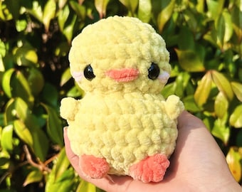 May include: A crocheted yellow duck plush toy with pink feet and black eyes. The duck is sitting in a person's hand.