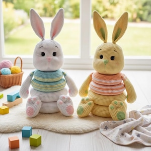 May include: Two crochet bunny stacking toys. One is white with blue and yellow rings, the other is yellow with orange and white rings. Both have long ears, black eyes, and pink accents. They sit on a white rug near a window.
