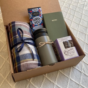 May include: A brown cardboard box filled with a cozy gift set. The box contains a plaid blanket, a travel mug, a small notebook with the word "NOTES" on the cover, a chocolate bar with the words "Thank You!" on the wrapper, and a box of lavender chocolate cookies.