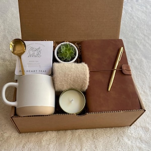 Pode incluir: Uma caixa de cartão castanho cheia de um conjunto de presente para autocuidado. O conjunto inclui uma caneca branca, uma pequena suculenta em vaso, um diário de couro castanho com uma caneta dourada, uma vela branca e um cachecol bege. A caixa também inclui um pequeno cartão com o texto "Minty White White Tea Big Heart Teas".