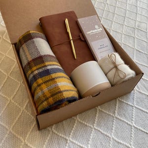 May include: A brown cardboard gift box filled with a plaid scarf, a brown leather journal with a gold pen, a white candle, a small white bag, and a chocolate bar with the text "moonstruck" on it.