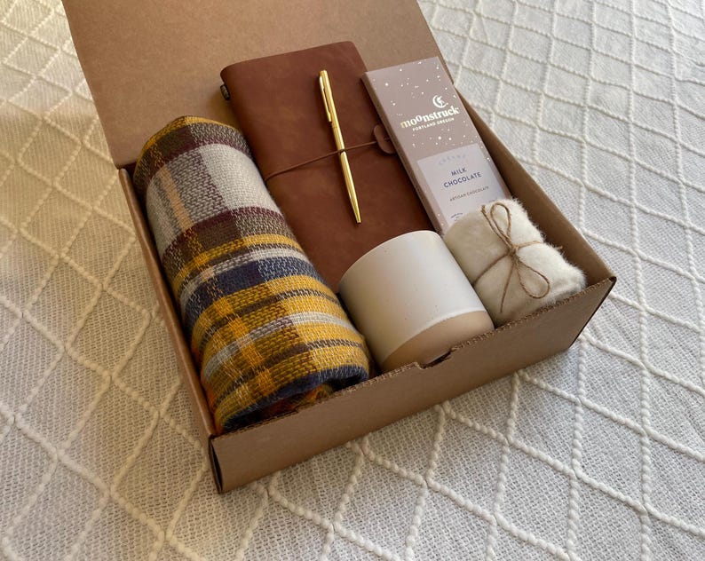 May include: A brown cardboard gift box filled with a plaid scarf, a brown leather journal with a gold pen, a white candle, a small white bag, and a chocolate bar with the text "moonstruck" on it.
