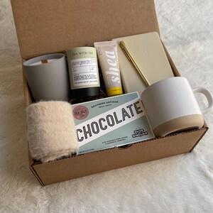 May include: A brown cardboard gift box filled with a variety of items, including a white mug, a candle, a hand cream, a notebook, a pen, and a small bag of chocolate. The box is open and the items are visible.
