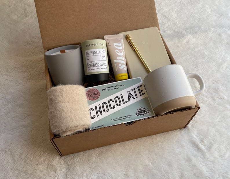 May include: A brown cardboard gift box filled with a variety of items, including a white mug, a candle, a hand cream, a notebook, a pen, and a small bag of chocolate. The box is open and the items are visible.