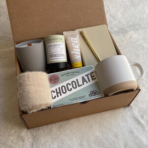 May include: A brown cardboard gift box filled with various items, including a white mug, a candle, a hand cream, a notebook, a pen, and a small bag of bath salts. The box also includes a card with the text "Southern Artisan Small Batch Chocolate".