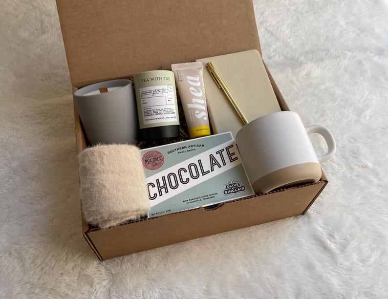 May include: A brown cardboard gift box filled with various items, including a white mug, a candle, a hand cream, a notebook, a pen, and a small bag of bath salts. The box also includes a card with the text "Southern Artisan Small Batch Chocolate".