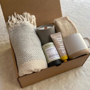 May include: A brown cardboard box filled with a self-care gift set. The set includes a gray candle, a white mug with a brown rim, a white and gray patterned throw blanket, a tube of shea butter lotion, and a small bottle of tea with the text "Tea with Tan" on the label.