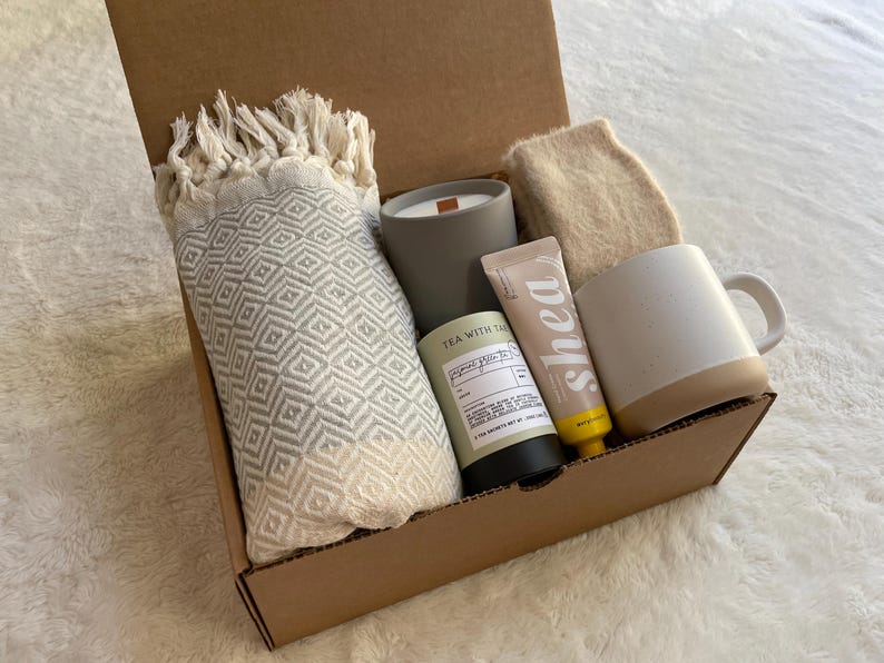 May include: A brown cardboard box filled with a self-care gift set. The set includes a gray candle, a white mug with a brown rim, a white and gray patterned throw blanket, a tube of shea butter lotion, and a small bottle of tea with the text "Tea with Tan" on the label.