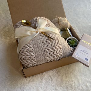 May include: A gift box filled with a cozy knitted throw blanket, a candle, a small succulent plant, and a bar of milk chocolate. The box is tied with a white satin ribbon and a gold spoon.