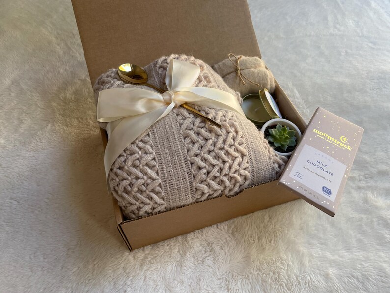 May include: A gift box filled with a cozy knitted throw blanket, a candle, a small succulent plant, and a bar of milk chocolate. The box is tied with a white satin ribbon and a gold spoon.