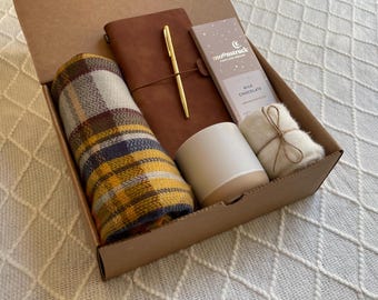 May include: A brown cardboard gift box filled with a variety of items, including a plaid scarf, a brown leather journal with a gold pen, a white candle, a bar of chocolate, and a small white fluffy item. The box is open and the items are visible.
