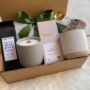 May include: A brown cardboard gift box filled with a gray candle, a bag of coffee labeled "Coffee Early Bird Blend", a bar of milk chocolate labeled "Moonstruck Milk Chocolate Artisan Chocolate", and a white ceramic mug with a brown bottom.