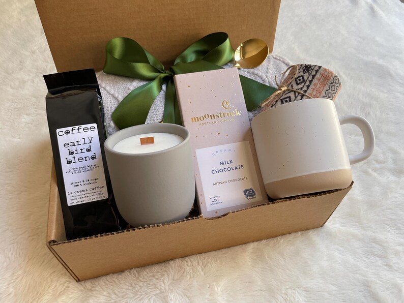 May include: A brown cardboard gift box filled with a gray candle, a bag of coffee labeled "Coffee Early Bird Blend", a bar of milk chocolate labeled "Moonstruck Milk Chocolate Artisan Chocolate", and a white ceramic mug with a brown bottom.