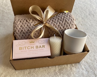 May include: A gift box filled with a cozy pink knitted throw blanket, a white ceramic mug, a small honeycomb candle, and a pink box with the text "Bitch Bar" and the words "Sweet Musk - Magnolia Blossom Fragrance".