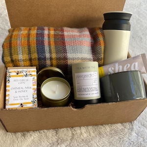 May include: A brown cardboard box filled with a cozy gift set. The set includes a plaid throw blanket, a candle, a bar of soap, a travel mug, and a tube of shea butter. The box is labeled "Red Circle Gifts" and "Tea with Tae".