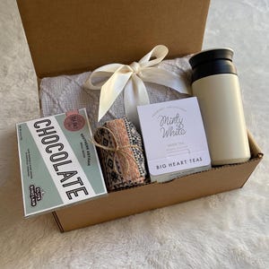May include: A brown cardboard gift box filled with a white blanket, a small box of chocolate, a tea bag packet, and a white insulated travel mug. The tea bag packet says "Minty White" and "Big Heart Tea".