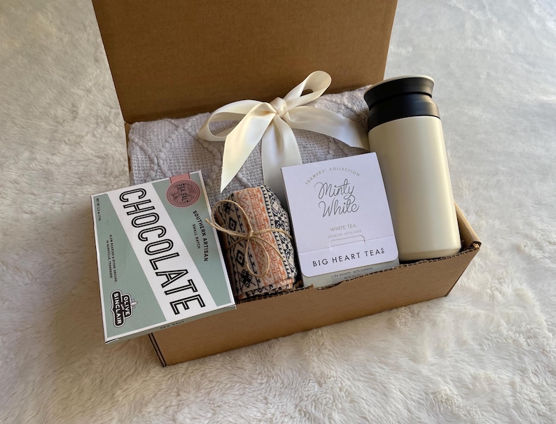 May include: A brown cardboard gift box filled with a white blanket, a small box of chocolate, a tea bag packet, and a white insulated travel mug. The tea bag packet says "Minty White" and "Big Heart Tea".