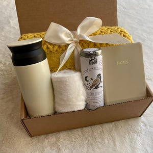 May include: A brown cardboard gift box filled with a white thermal bottle, a white knitted scarf, a small tin of tea with the text "Dream Catcher" and a cream-colored notebook with the word "NOTES" on the cover.