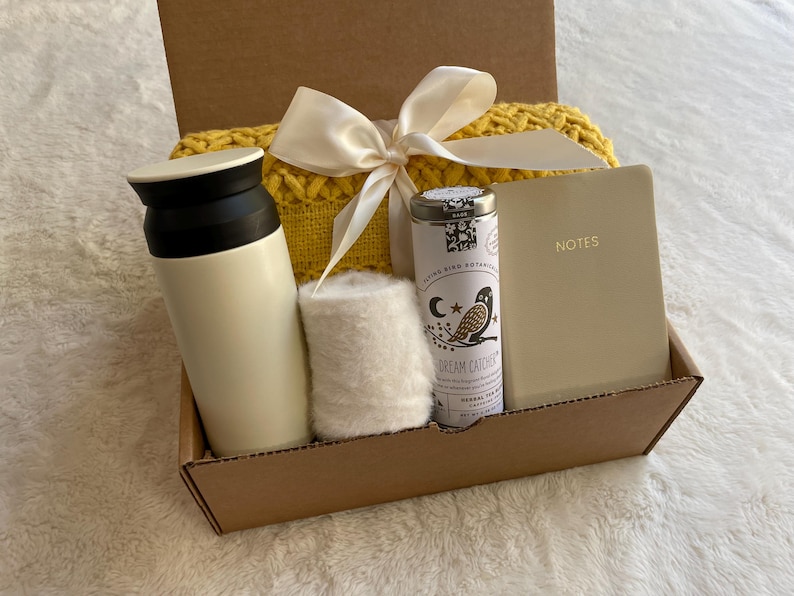 May include: A brown cardboard gift box filled with a white thermal bottle, a white knitted scarf, a small tin of tea with the text "Dream Catcher" and a cream-colored notebook with the word "NOTES" on the cover.