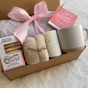 May include: A brown cardboard gift box filled with a variety of items, including a white mug, a candle, a small package of cookies, a knitted throw blanket, and a small package of tea. The box is tied with a pink ribbon.