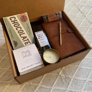 Puede incluir: Una caja de cartón marrón llena de un set de regalo. El set incluye una barra de chocolate, un pequeño frasco de café, una vela, una bolsita de té y un diario encuadernado en cuero con una pluma dorada.