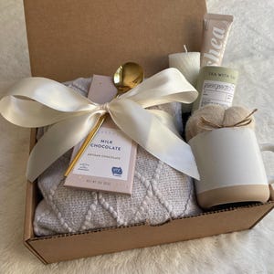 May include: A brown cardboard gift box filled with a cozy white blanket, a mug, a bar of milk chocolate, a gold spoon, and a candle. The box is tied with a white satin ribbon.