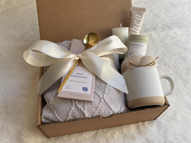 May include: A brown cardboard gift box filled with a cozy white blanket, a mug, a bar of milk chocolate, a gold spoon, and a candle. The box is tied with a white satin ribbon.