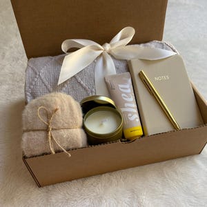 May include: A brown cardboard box filled with a self-care gift set. The box contains a white throw blanket, a small candle, a tube of shea butter lotion, a small notebook with the word "NOTES" on the cover, and a gold pen.