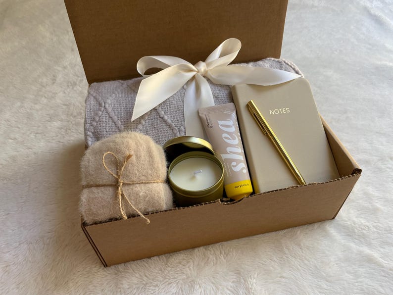 May include: A brown cardboard box filled with a self-care gift set. The box contains a white throw blanket, a small candle, a tube of shea butter lotion, a small notebook with the word "NOTES" on the cover, and a gold pen.