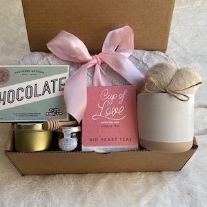 May include: A gift box filled with a variety of items, including a bar of chocolate, a jar of honey, a small jar of tea, a mug, a tea bag, and a knitted blanket. The box is decorated with a pink ribbon and the text "Cup of Love" is visible on the tea bag.