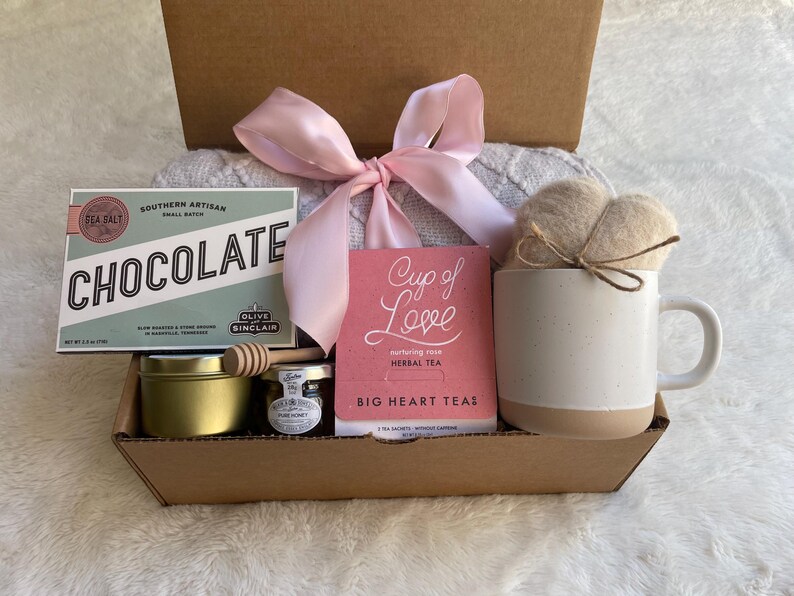 May include: A gift box filled with a variety of items, including a bar of chocolate, a jar of honey, a small jar of tea, a mug, a tea bag, and a knitted blanket. The box is decorated with a pink ribbon and the text "Cup of Love" is visible on the tea bag.