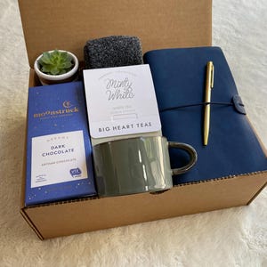 May include: A brown cardboard box filled with a variety of gifts, including a mug, a bar of dark chocolate, a small potted succulent, a notebook, a pen, and a package of tea. The tea package has the text "Minty White" and "BIG HEART TEAS". The chocolate bar has the text "DREAM DARK CHOCOLATE" and "MOONSTRUCK".