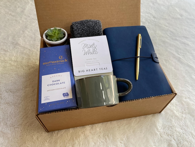 May include: A brown cardboard box filled with a variety of gifts, including a mug, a bar of dark chocolate, a small potted succulent, a notebook, a pen, and a package of tea. The tea package has the text "Minty White" and "BIG HEART TEAS". The chocolate bar has the text "DREAM DARK CHOCOLATE" and "MOONSTRUCK".