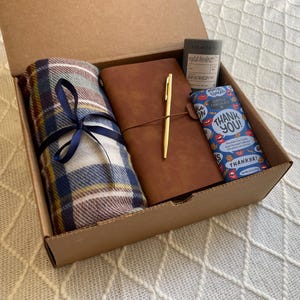 May include: A brown cardboard box filled with a gift set. The set includes a plaid blanket, a brown leather journal with a gold pen, and a blue and white chocolate bar with the text "Thank You!" printed on it.