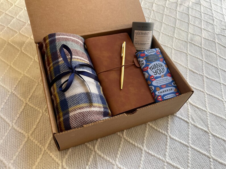 May include: A brown cardboard box filled with a gift set. The set includes a plaid blanket, a brown leather journal with a gold pen, and a blue and white chocolate bar with the text "Thank You!" printed on it.