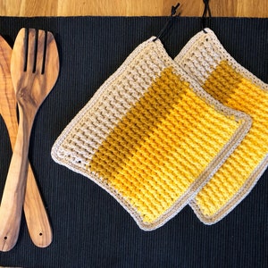 Könnte beinhalten: Zwei gehäkelte Topflappen mit einem beige-gelben Farbverlauf. Die Topflappen liegen auf einer schwarzen Oberfläche mit zwei hölzernen Küchenutensilien.