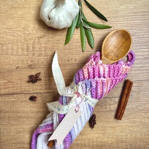 Könnte beinhalten: Ein handgefertigter, gestrickter Küchenutensilienhalter in Rosa-, Lila- und Weißtönen, der einen Holzlöffel hält. Der Halter ist mit einem Band und einem Etikett mit der Aufschrift "KLEINIGKEIT" versehen. Knoblauch, Rosmarin, Sternanis und ein Zimtstange sind ebenfalls vorhanden.