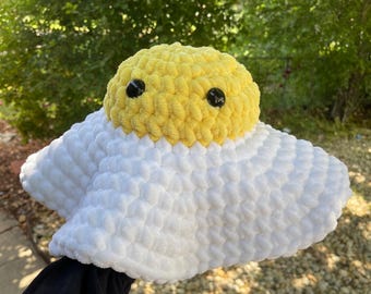 Puede incluir: Un juguete de peluche hecho a mano con forma de huevo tejido a ganchillo. La yema es amarilla con dos ojos negros de botón, y el blanco tiene una forma esponjosa, como una nube. El juguete se sostiene contra un fondo de follaje verde.
