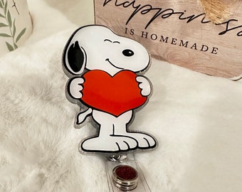 May include: A Snoopy-themed retractable badge reel. The design features Snoopy holding a red heart. The reel has a clear plastic strap and a glittery red button. The background includes a wooden sign that says "happiness is homemade."