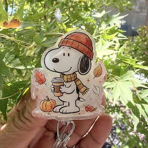May include: A clear acrylic badge reel featuring a cartoon illustration of Snoopy wearing an orange knit beanie and a yellow and brown striped scarf. The character holds a coffee cup next to a small orange pumpkin and scattered autumn leaves.