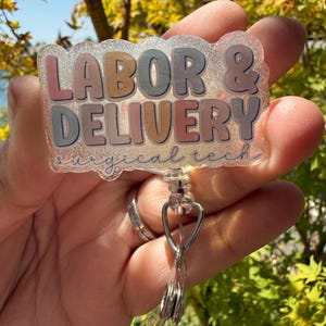 May include: A clear, glittery badge reel with the words "LABOR & DELIVERY" in pastel colours. The words "surgical tech" are printed below. The badge reel has a silver metal clip and a clear plastic retractable cord.