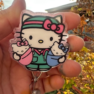 May include: A pink glitter badge reel featuring a Hello Kitty design dressed as a nurse, holding two babies. The design includes a pink bow, green scrubs, and a retractable clip.
