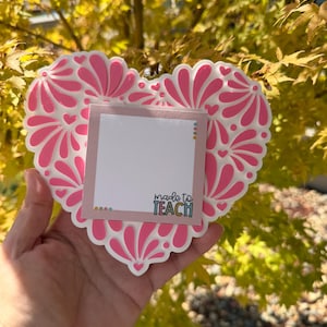 Puede incluir: Un objeto decorativo en forma de corazón, blanco y rosa, con un diseño floral. Se adjunta un pequeño bloc de notas cuadrado, con las palabras "made to TEACH" impresas. El objeto se sostiene frente a un fondo de hojas amarillas y verdes.