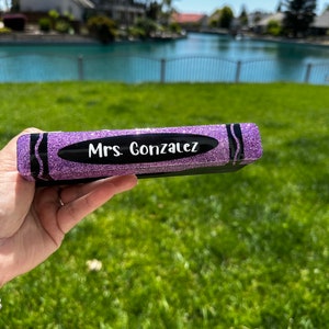 Puede incluir: Una caja con forma de crayón de purpurina morada con el nombre "Mrs. Gonzalez" en letras blancas. La caja tiene detalles en negro. Se sostiene sobre un fondo de hierba verde, un lago y casas.