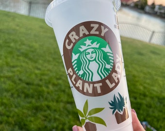 May include: A clear plastic reusable cold beverage cup with a lid. The cup features a green and brown logo with the words "CRAZY PLANT LADY" and plant illustrations. The cup is held in front of a green lawn and a body of water.