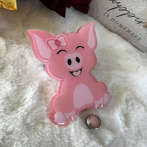 Puede incluir: Un portatarjetas en forma de cerdo rosa con una cara sonriente y un lazo. El portatarjetas tiene un cordón retráctil de plástico transparente y un botón decorativo con purpurina. El cerdo tiene un cuerpo rosa claro y un contorno rosa más oscuro.