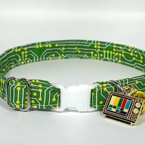 May include: Green cat collar featuring a circuit board design and a white plastic buckle. The collar has a gold-coloured bell and a charm shaped like a retro television set.