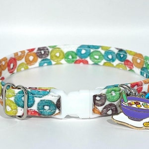 May include: A white cat collar with a colorful cereal print and a charm of a bowl of cereal with a spoon. The charm is attached to a silver bell.